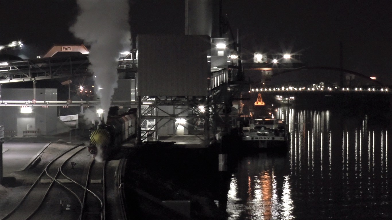 Bei Nacht rangiert eine Dampfspeicherlokomotive eine Wagenreihe an einer hell
                  beleuchteten Umschlaganlage am Wasser. Im Hintergrund Kräne, ein Frachtschiff
                  und eine beleuchtete Bogenbrücke, deren Lichter sich im Fluss spiegeln.