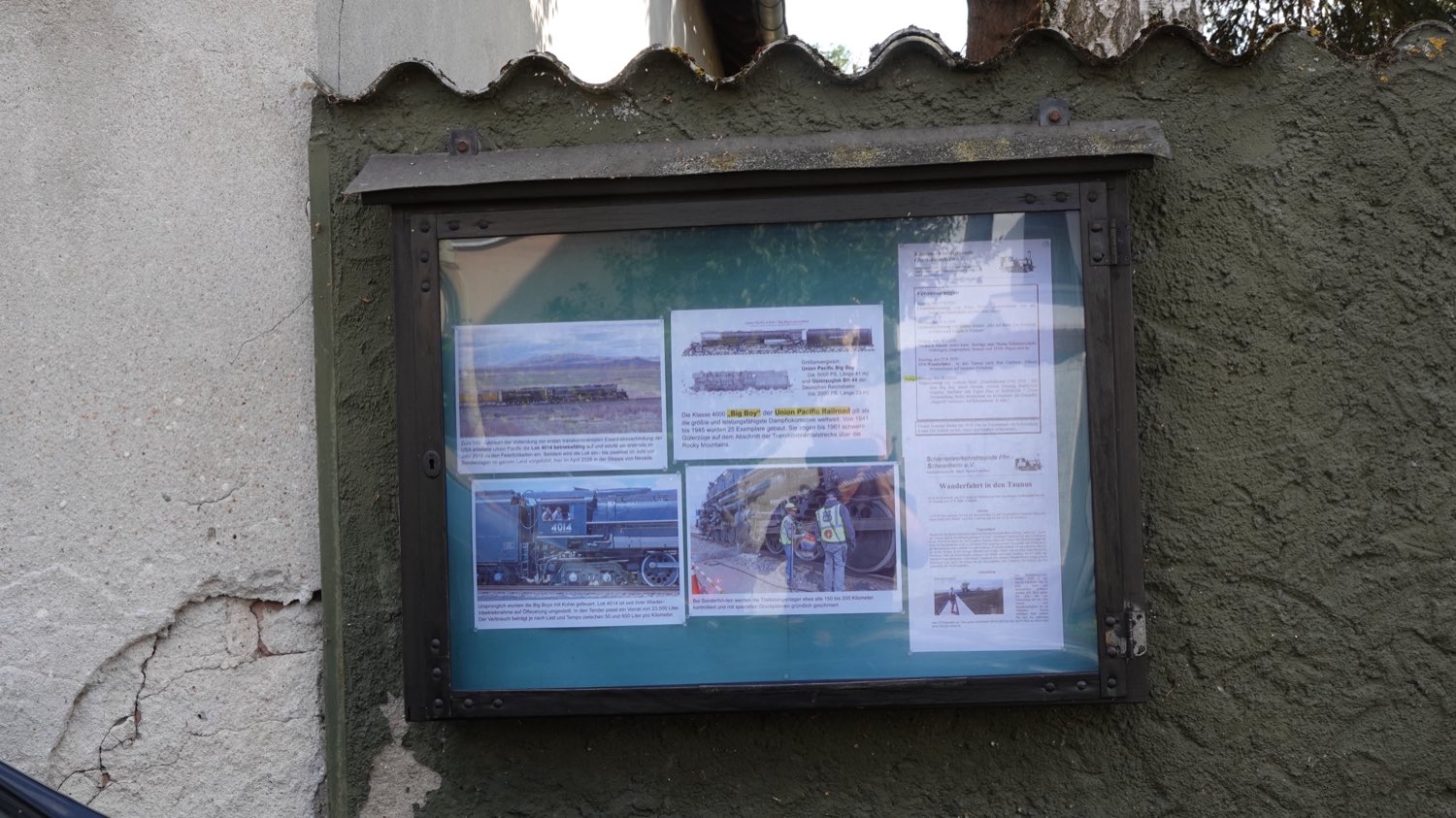Ein hölzerner Schaukasten mit Blechdach an einer verputzten Hauswand.
                    Hinter der Scheibe hängen Fotos und Texte über die Dampflokomotive
                    „Big Boy“ der Union Pacific, daneben das Veranstaltungsprogramm des
                    Vereins.