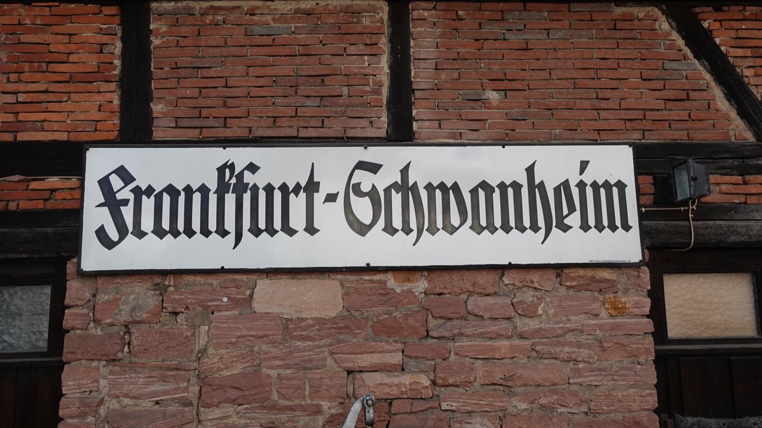 Ein emailliertes Stationsschild mit der Aufschrift „Frankfurt-Schwanheim“
                      in Frakturschrift, angebracht an einer Fachwerkwand aus Backstein und
                      rotem Sandstein.