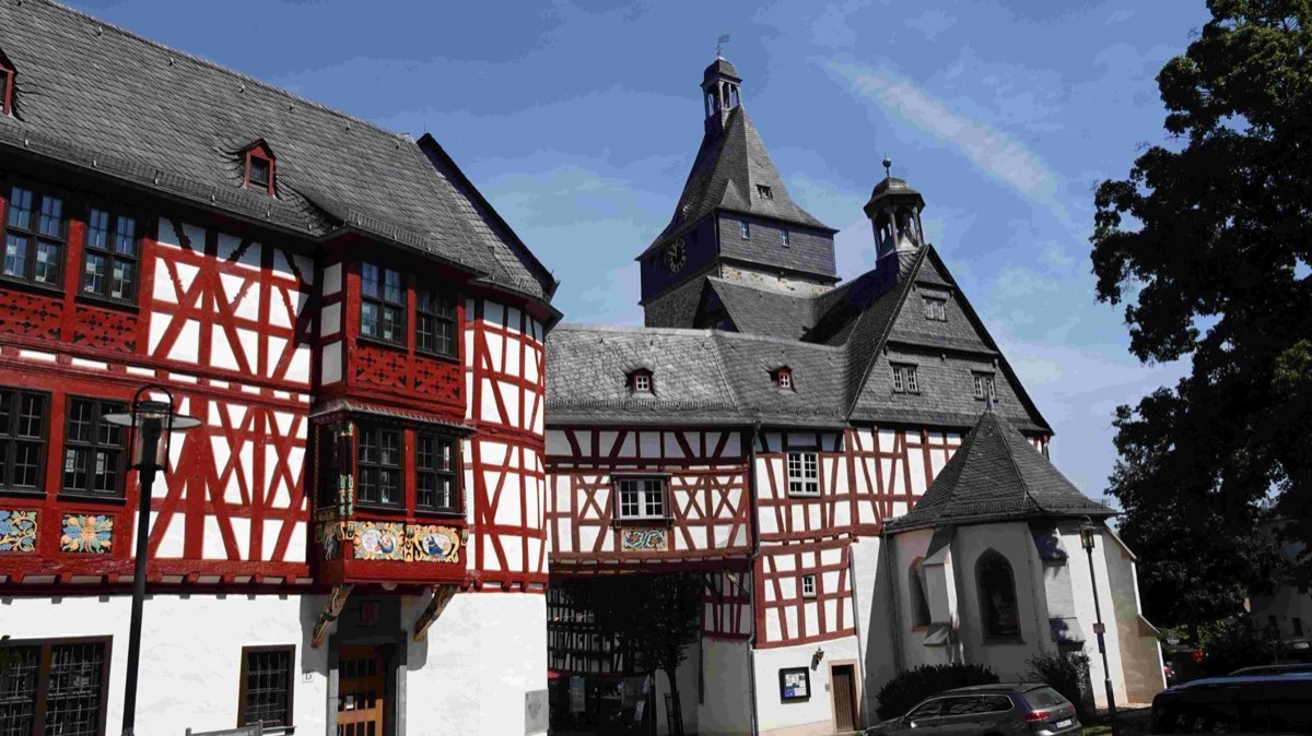 Ein großes Fachwerkhaus mit rotem Gebälk, bemalten Brüstungsfeldern und
                      einem Erker. Dahinter ein hoher Torturm mit geschweifter Haube und eine
                      kleine weiße Kapelle.