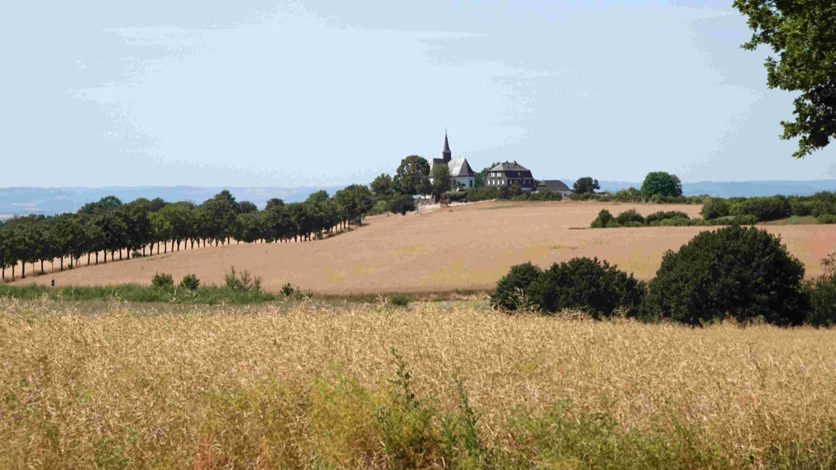 Hinter abgeernteten Feldern und einer Baumreihe steht auf einer Anhöhe eine
                      weiße Kapelle mit spitzem Turm, daneben ein größeres Haus.