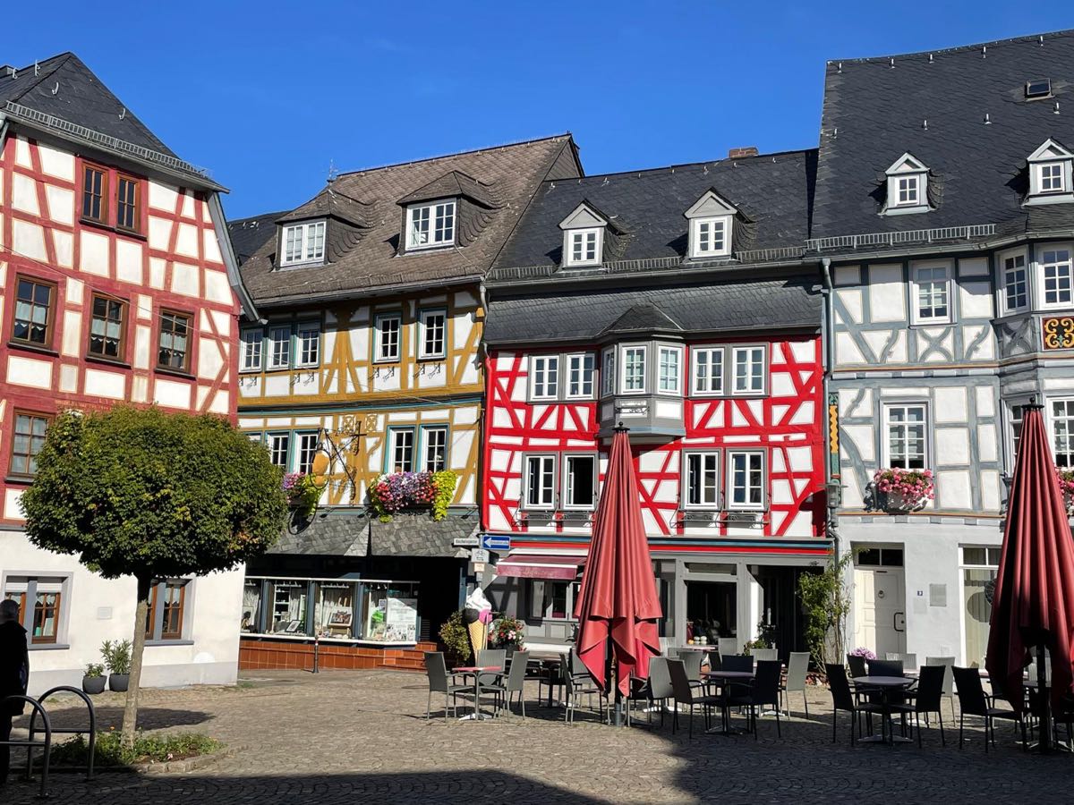 Eine Reihe bunter Fachwerkhäuser in Rot, Ocker und Grau an einem
                      gepflasterten Platz, davor Tische, Stühle und zusammengeklappte
                      Sonnenschirme eines Cafés.