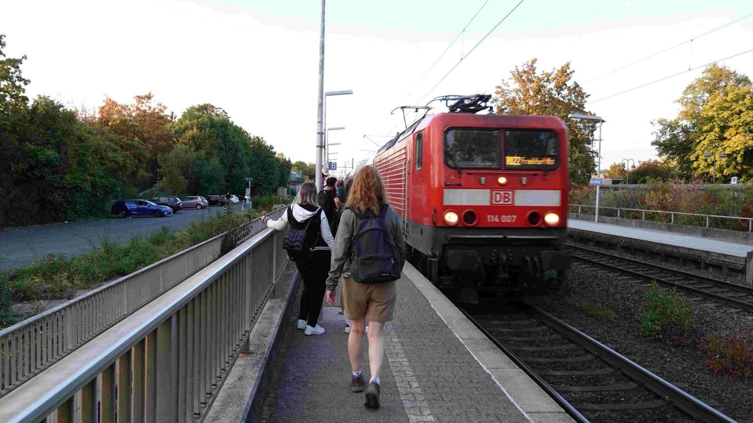 Am Bahnsteig in Bad Camberg fährt eine rote Elektrolok der Deutschen Bahn mit
                    der Nummer 114 007 ein. Zwei Mitreisende mit Rucksäcken gehen von hinten
                    gesehen auf den Zug zu.