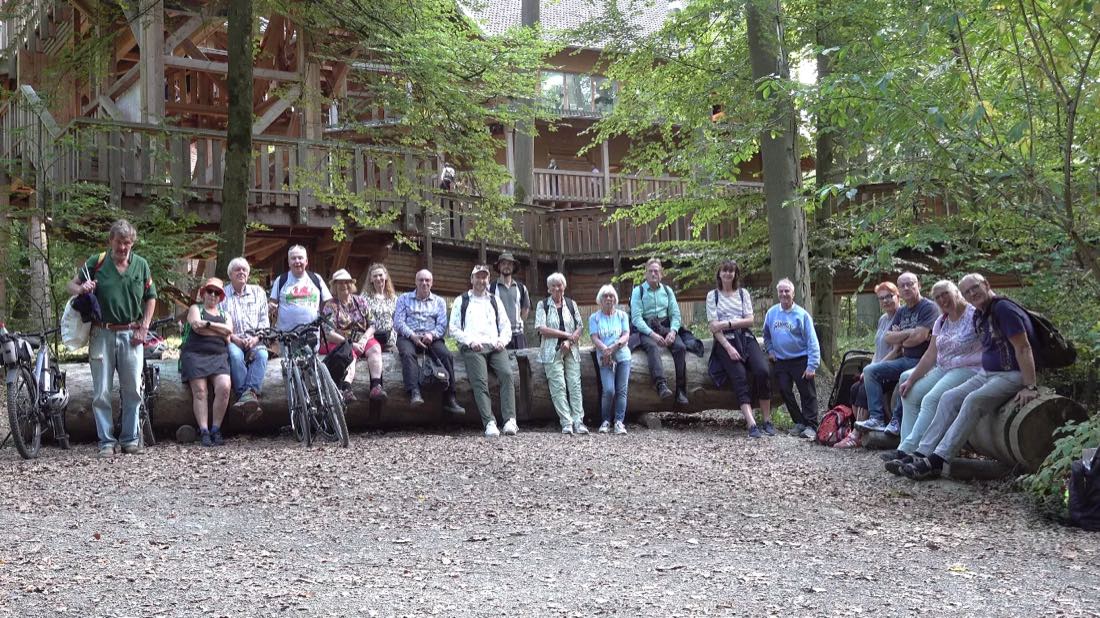 Eine Wandergruppe sitzt und steht nebeneinander auf einem liegenden
                    Baumstamm im Wald, davor mehrere Fahrräder. Dahinter ein großer Holzbau mit
                    umlaufenden Galerien zwischen hohen Buchen.