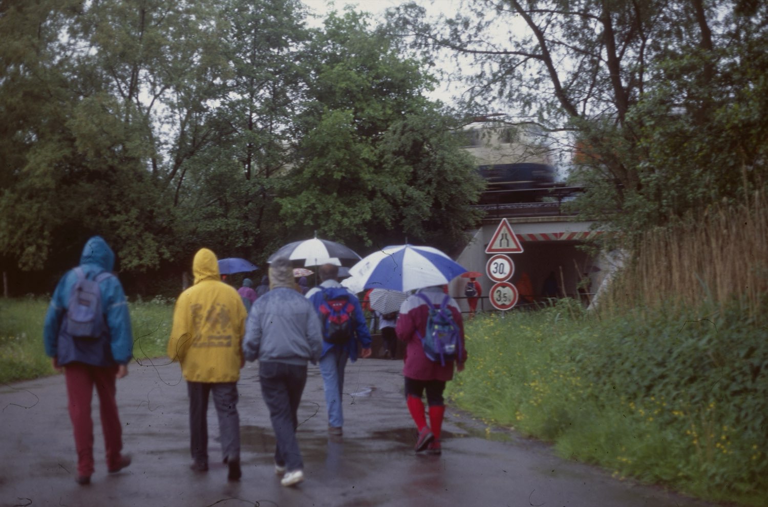 Eine Wandergruppe in Regenjacken und mit aufgespannten Schirmen geht auf
                    einem nassen Weg auf eine Eisenbahnunterführung zu; oben auf der Brücke
                    fährt ein Zug vorbei.