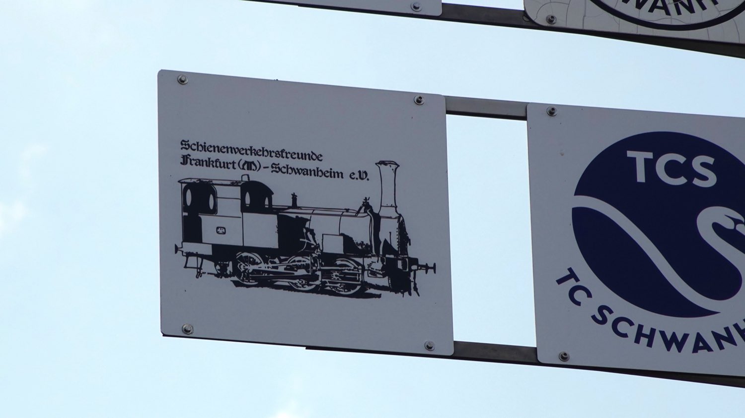 Ein weißes Emailschild am Mast des Wappenbaums vor blauem Himmel: die
                      Dampflok des Vereinszeichens, darüber in Frakturschrift
                      „Schienenverkehrsfreunde Frankfurt (M)-Schwanheim e. V.“ Daneben das
                      Schild des Tennisclubs Schwanheim.